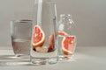 Glass carafe and bottle with grapefruit slices in water on a neutral background