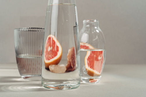 Glass carafe and bottle with grapefruit slices in water on a neutral background